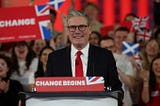 Keir Starmer stands behind a podium labelled ‘CHANGE BEGINS’, behind him a Labour supports holds up a sign reading ‘CHANGE’
