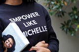 Woman wears navy sweatshirt with words “Empower Like Michelle”. She carries Michelle Obama’s book “Becoming” under her arm.