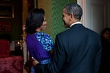 Barack and Michelle Obama are standing close together and talking in the White House Green Room. Michelle wears a patterned skirt and a dark top.  Barack wears a dark suit.
