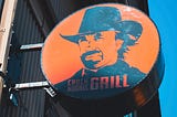 An orange sign featuring Chuck Norris in a cowboy hat. The sign reads, “Chuck Norris Grill.”