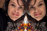 Ariel, a white woman in her late ’40s, leans her face against a mirror. She and her reflection are both looking into the lens of the camera and smiling. There are flowers in the background.