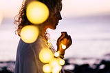 A woman, who is just out of focus, clutches a string of lights on a twilight beach.