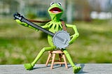 Kermit the Frog sits on a stool and plays the banjo