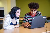 Two young people sitting at a table look at a laptop together.