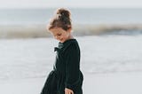 A young child (girl) standing on a beach smiling and looking down at the sand.