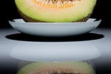 A close-up image of a cantaloupe or honeydew melon slice placed on a white plate against a black background. The melon has pale green flesh with seeds still attached to the top. Its reflection is visible on the glossy surface beneath, creating a mirrored effect that enhances the overall composition. The lighting highlights the texture of the fruit and contrasts with the dark surroundings.