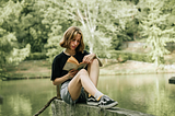 A blonde woman in a black t-shirt and shorts reads a book outside