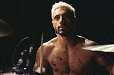 Rueben (played my Riz Ahmed) sits shirtless at his drum set in a dimly-lit room.
