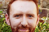 Photo is a headshot of a man with short red hair and a beard looking directly at the camera with a slight smile