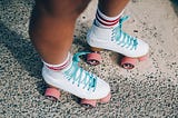 A rollerskater’s legs from the knees down — white roller skates, striped white and red socks, pink wheels.