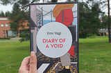 Fariza’s hand holding the copy of Diary of a Void with trees on grass and a building in the background