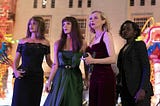 Four women dressed to the nines stand outside a lush building and look off into the distance. From left, they are a brunette in a black off-the-shoulder evening dress, a long-haired brunette in a green evening gown, a blonde in a black top with thin straps and chic slacks, and a black woman with chin-length hair dressed in a sharp black blazer and slacks.