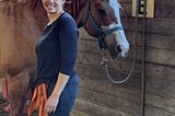 Young woman with her hair in a ponytail, wearing a blue shirt and jeans, stands smiling in front of a horse.