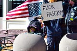 Protesters hold the U.S. flag upside down as a symbol of constitutional emergency, along with a sign that reads “Fuck Fascism”