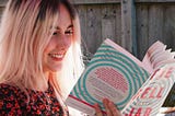 Photo of myself, sat outside in the sunshine reading a copy of The Bell Jar with a swirling aqua and pink cover.