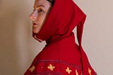 A red medieval knit hood that drapes as a collar around the shoulders with yellow four-point flowers on the border. It is being worn by a white female looking to the side against a plain white backdrop.