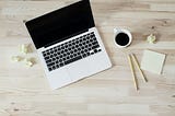 A laptop, coffee, and notepad on a wooden desk.