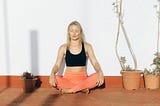 Woman in coral exercise pants sitting with her legs crossed, hands on her knees, and eyes closed.