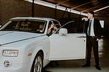 A woman sits inside a rolls-royce, and a man in a suit stands by the car door