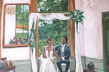 landscape format detail from live wedding painting. Bride and groom sitting indoors in front of a backdrop and laughing to each other. In fron of mirror and window. Outside lots of greens.