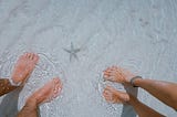 Feet of man and woman in shallow beach water with starfish between them on white sand.