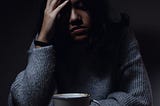 An image of a young woman in seated in a dark setting with head held against her right palm, in a depressed pose.