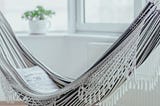 A grey and white fringed hammock with pillow inside a light-filled room with windows, a plant in a white vase.