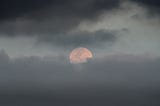 a pink moon at twilight in a cloudy sky.