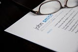 Photo displays a resume laying on a dark surface. The top of the resume has the name “John Smith” and there are glasses laid on the top of the resume.