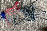 A variety of different insulated electrical wires on a concrete floor. Red, green, blue, and grey. The insulation is stripped of the end of each one.