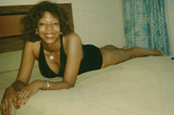 Picture of a smiling light skinned Black young woman laying width-wise across a made up bed wearing black summer shorts and sleeveless top.