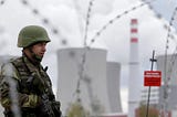 A military guard, framed by razor wire, stands on watch outside a power station, with cooling towers in the background