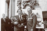 black and white photo of Arnie Huber holding his own bust and posing for a photographi with a man in a suit during Huber’s Hall of Fame induction ceremony.