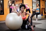 Young ethnic woman with fit ball sitting on floor while training in modern gym