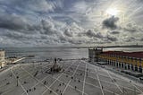 A wide-angle view of a large public square in Lisbon, Portugal, called Praça do Comércio. The square is paved with geometric patterns and has a central statue surrounded by people walking. On the right, there are buildings with yellow facades and a red roof, and in the background, a body of water stretches towards the horizon under a dramatic cloudy sky, with the sun peeking through.