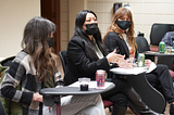 Three women wearing masks sit at desks engaged in conversation. An Asian American woman with a black mask, long black hair and a black blazer gestures with her hands while she speaks. A woman with long red hair and a black mask looks on from one side, listening. A woman with long brunette hair, a black mask and a checked wool shirt looks on from the other side.