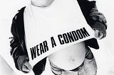 Black and white photo of Sinead O’Connor in a jacket and jeans, wearing a white shirt with WEAR A CONDOM printed on it. By Kate Garner in 1986.