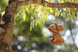 An image of a tree branch in the sunlight. From the branch hang three small, light-brown ceramic prayer bells. The bells hang from a thin string in a bunch, catching the sunlight along their tops.