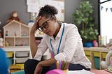 A young Black teacher sits in a preschool classroom with herhand on her head. Photo by Krakenimages.com/Adobe Stock