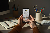 Hands holding a phone showing the Ladies Who Design TikTok profile, with art supplies and a sketchbook on the desk in soft afternoon light