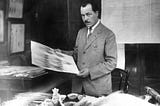 A man, Nikolai Vavilov, stands over a desk in a suit, holding up a scientific print. His desk is covered in plant samples