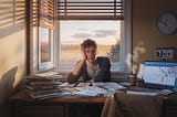 A young adult sits at a cluttered desk by a window, looking stressed in the early morning light, with a clock showing 7:00 AM.