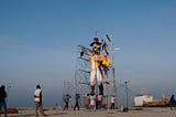 A group of people are mounting a huge effigy with ropes and scaffolding with a blue sky in the background.