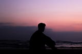 A dark masculine-presenting silhouette of someone sitting against a moody pink-purple sky.