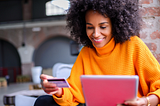 A woman sits by a window. She holds a credit card in her right hand and a tablet in her left hand.