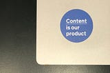 A blue sticker that says ‘Content is our product’ on a silver laptop