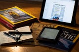 Display of magazines and newspapers, in print and digital, along with a Kindle and a pocket notebook and pens on a desk.