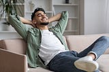 A young Arab man leaning back on a sofa, airpods in his ears, with his hands behind his head and a smile on his face. He is clearly enjoying spending this time by himself.