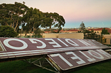 A vintage sign for Hotel San Diego lays on the ground upside down. The city and trees are in the background.
