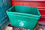 Two green recycling bins stacked on inside the other are placed against a red exterior wall and a green door. The bottom bin appears worn with a message encouraging recycling and an emblem representing sustainability.